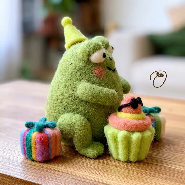 A green felt monster plush toy wearing a yellow party hat, sitting on a wooden table and hugging a small felt cupcake with pink and yellow frosting. A small rainbow gift box sits beside it, with a blurred indoor background.