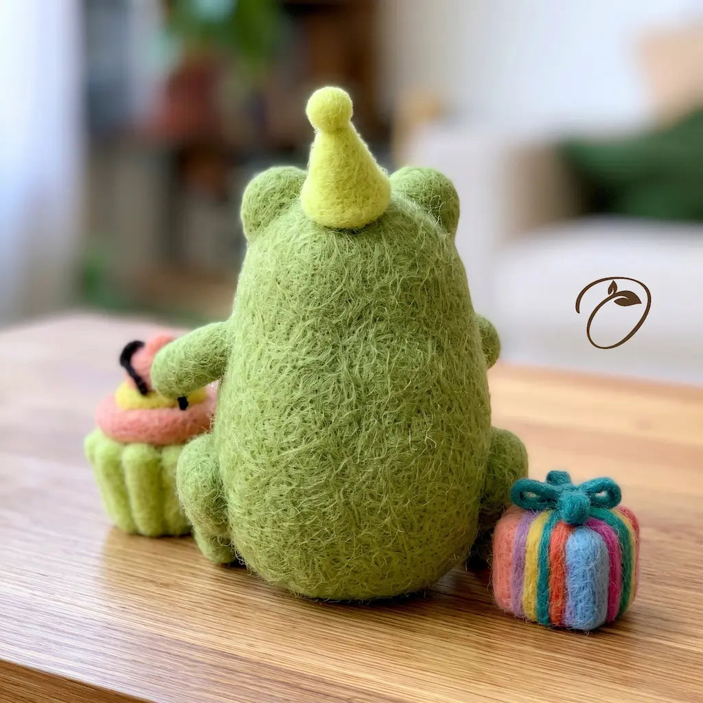 A cute green felt monster plush toy with a yellow party hat, sitting on a wooden table next to a small felt cupcake and a striped felt gift box.