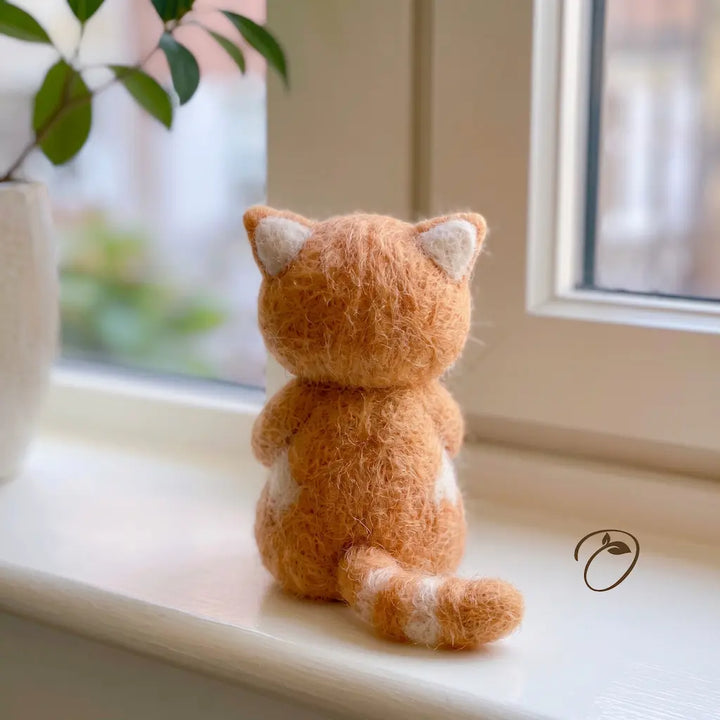 A small felted wool cat toy sits on a white windowsill, facing a window. The toy is made of soft orange-brown wool with white inner ears and white accents on its belly and tail.