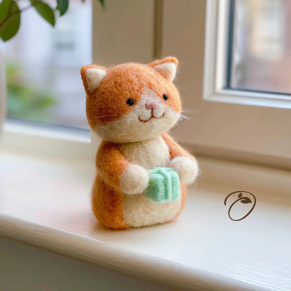 A small felted wool cat figurine sits on a white windowsill. The cat is orange and cream with a mint green soap dish in its paws.