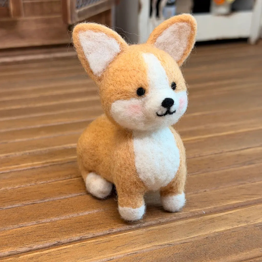 A small felt dog figurine resembling a corgi, with tan and white fur, black eyes, and a black nose, sitting on a wooden floor.