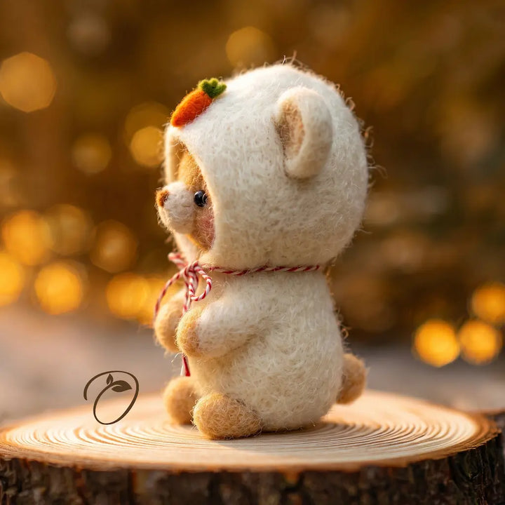 A small felt teddy bear wearing a cream hooded outfit with a carrot accent on its head, sitting on a round wood slice with a warm golden bokeh background.