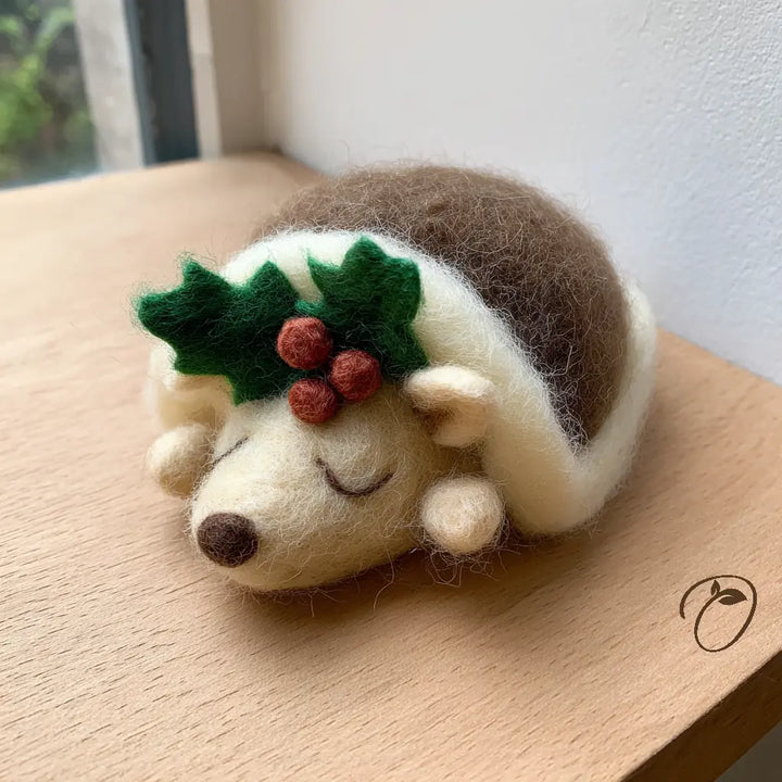 Sleeping felt hedgehog with closed eyes, cream trim, green holly leaves and three red berries on a wooden surface.
