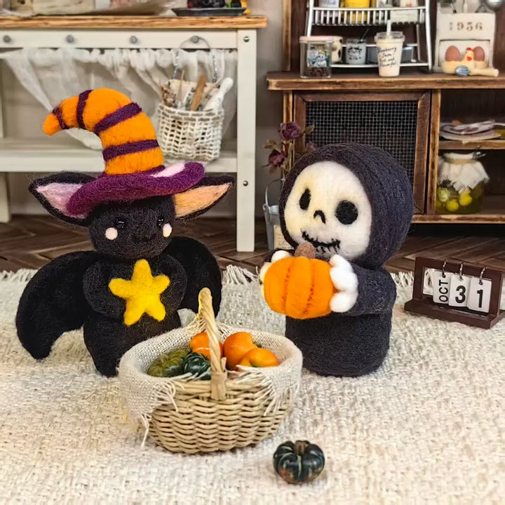 Two handmade felt wool Halloween plush toys in a cozy miniature room setting. One is a black bat wearing an orange and purple striped witch hat and holding a yellow star, next to a small woven basket of pumpkins.