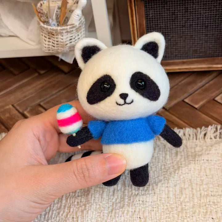 A small felt panda toy being held by a hand. The panda has a white body with black facial features and limbs, wearing a bright blue shirt, and is holding a small striped popsicle accessory.