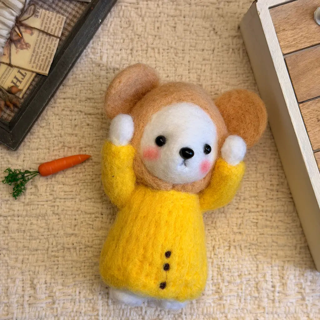 A small felted wool animal toy with a tan and white face, pink cheeks, and a bright yellow knitted body, posed on a textured beige fabric surface with small decorative props nearby.