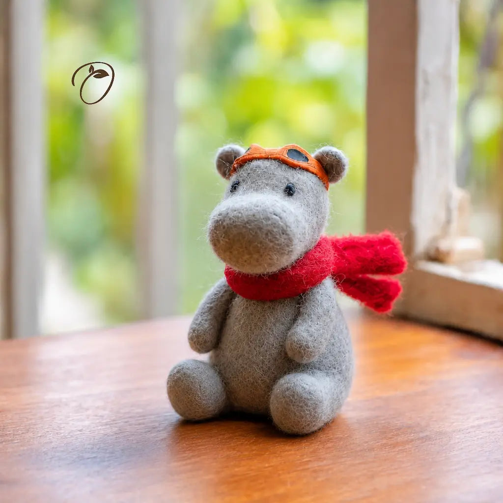Gray plush toy hippo wearing an orange hat and red scarf on a wooden surface with a blurred green background
