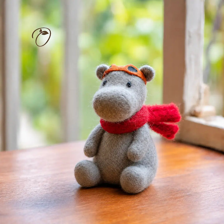 Gray plush toy hippo wearing an orange hat and red scarf on a wooden surface with a blurred green background
