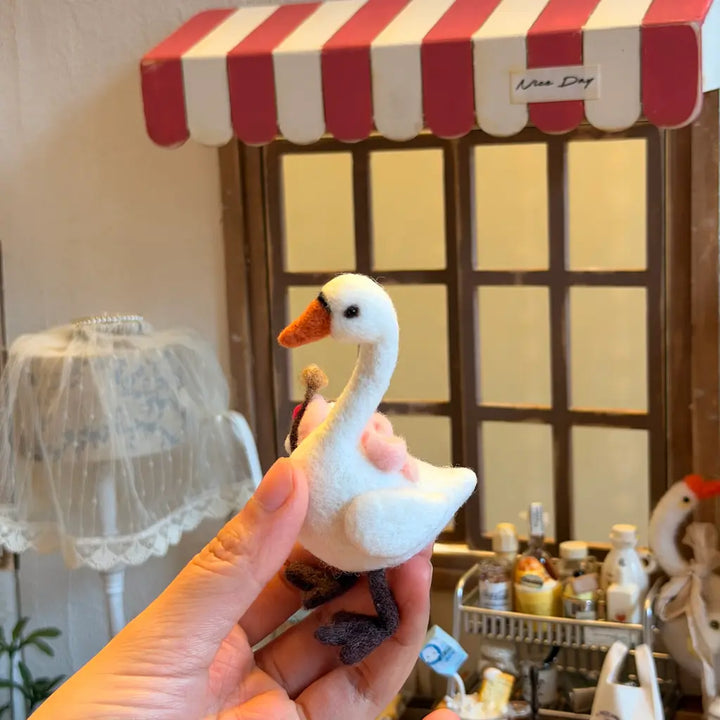 A hand holds a small felt goose with a white body, orange beak, and dark legs in front of a miniature shop window with a red-and-white awning.