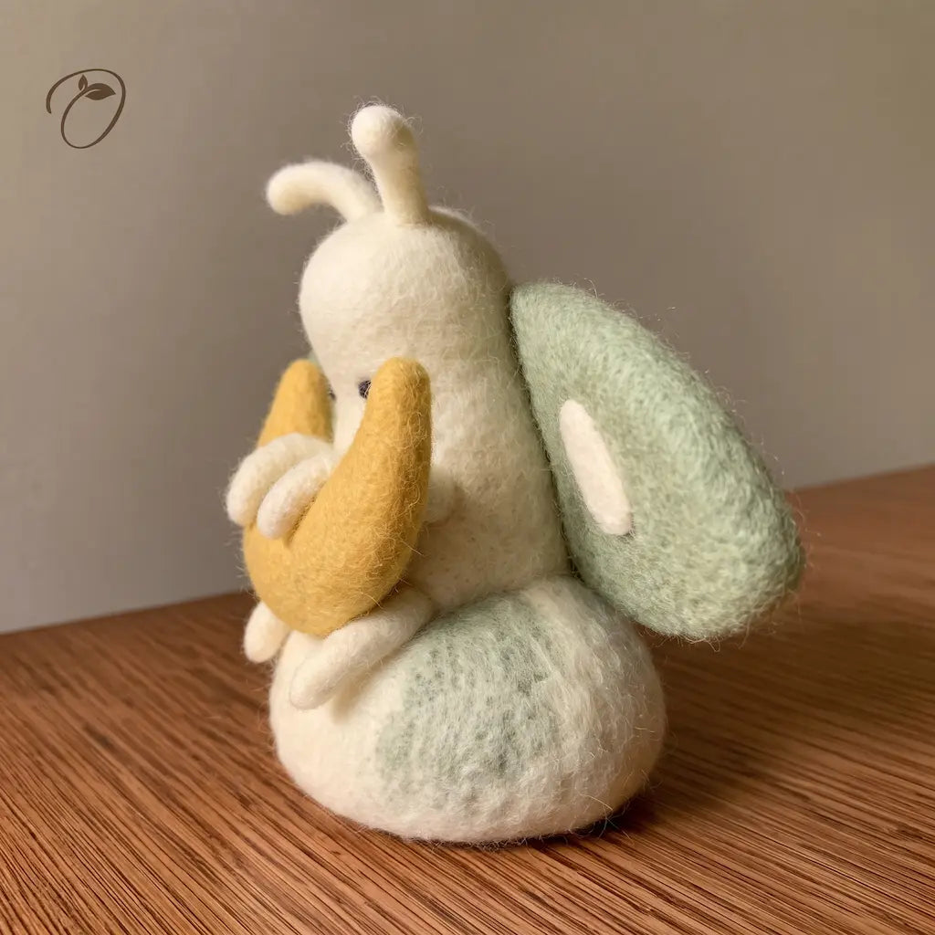 Side view of a wool felt moth cradling a golden crescent moon, showing its rounded ivory body and sage-green wing.