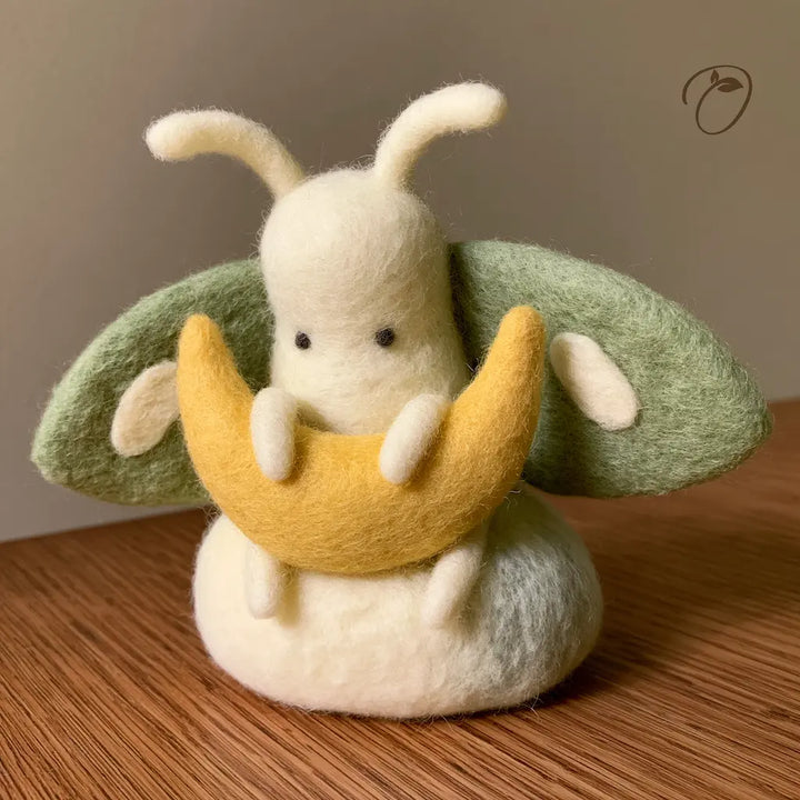 Ivory wool felt moth holding a golden crescent moon, with sage-green wings, pale oval markings and curved antennae.