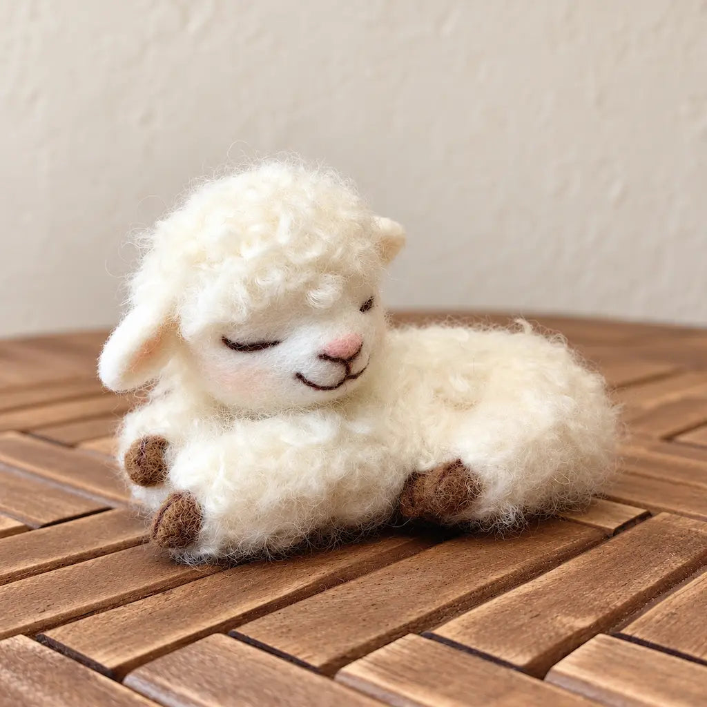Three-quarter view of a sleeping wool felt lamb showing its pink nose, tucked front paws, and thick cream-colored curly fleece.
