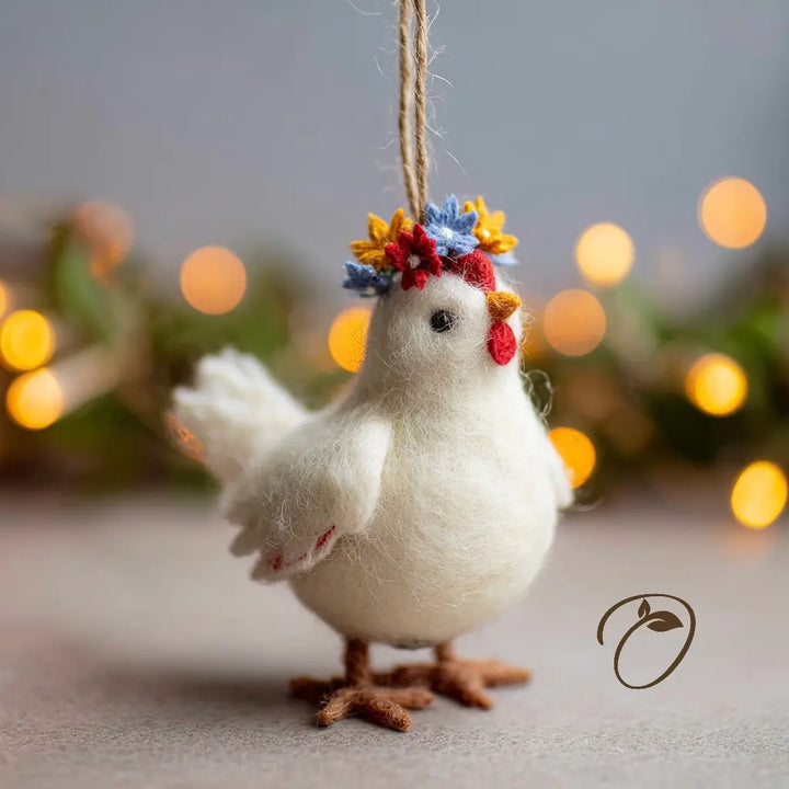 A white felt chicken ornament hanging by a twine string, decorated with a small multicolor floral crown and red comb detail, set against a softly blurred background with warm bokeh lights.