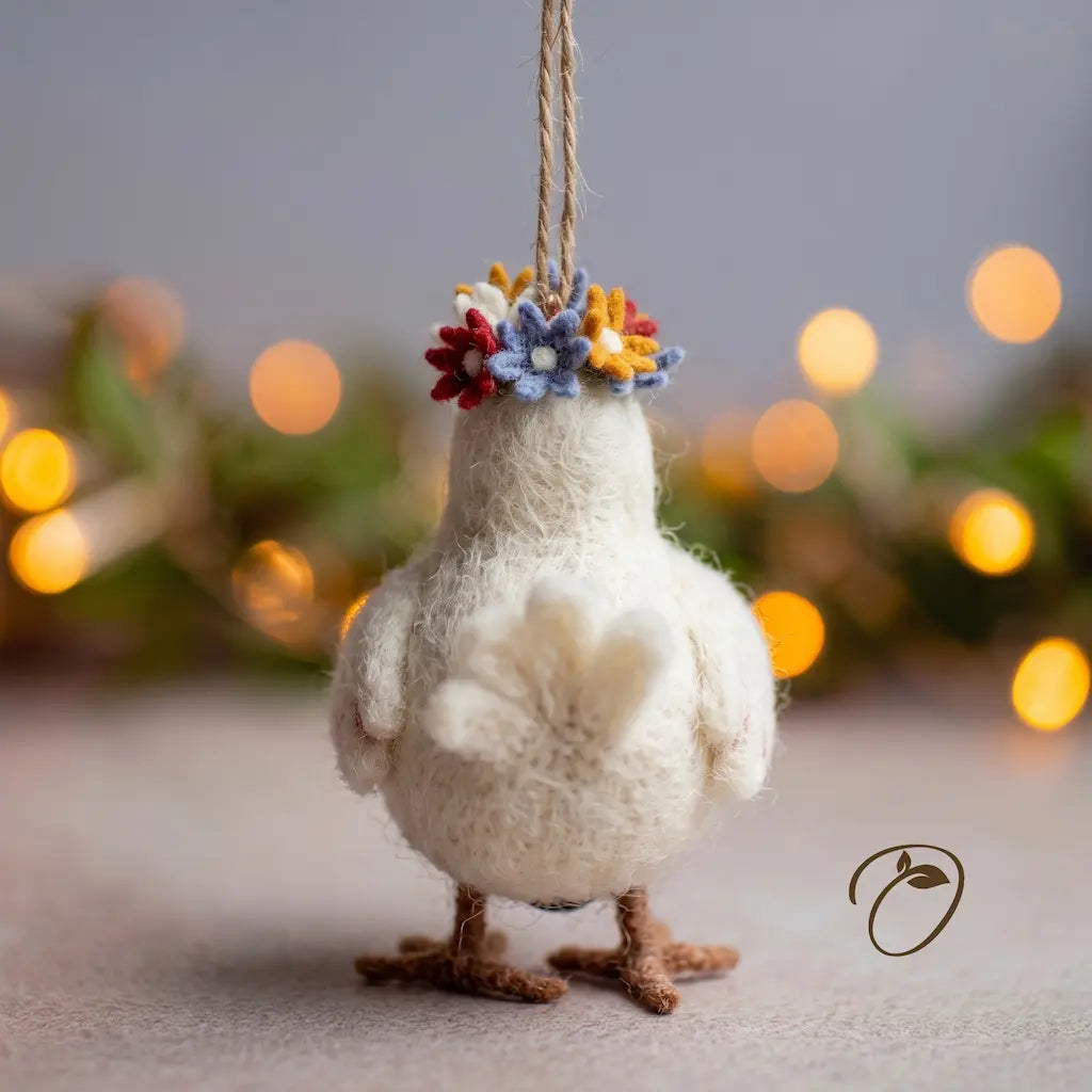 A small white felted wool bird ornament hanging by a twine string, with a colorful flower crown in red, yellow, blue, and white, set against a softly blurred background with warm bokeh lights.