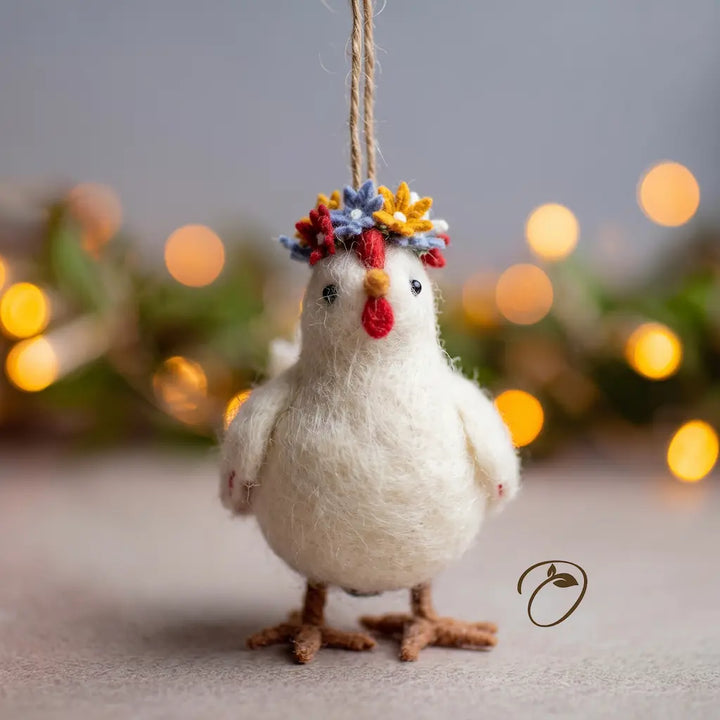 A white felt chicken ornament hanging by a twine string, with a small multicolor flower crown and red comb, photographed against a softly blurred warm bokeh background.