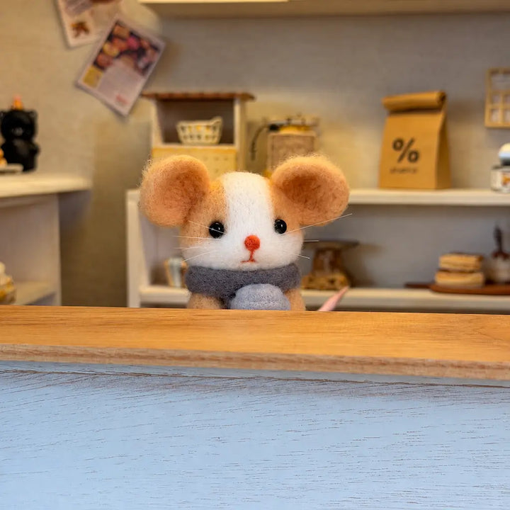 Felt mouse in a gray scarf peeking over a wooden café counter, with miniature shelves and a coffee bag behind it.