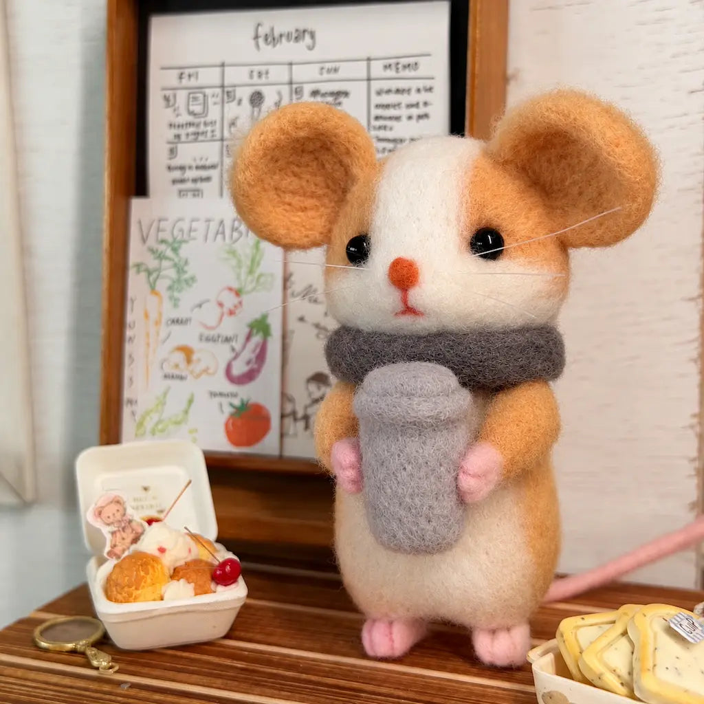 Front view of a felt mouse holding a gray coffee cup, with round tan ears, white whiskers and a pink tail.
