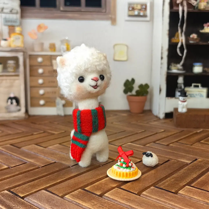 Cream wool felt alpaca wearing a red-and-green striped scarf on a miniature wooden floor beside small holiday food props.
