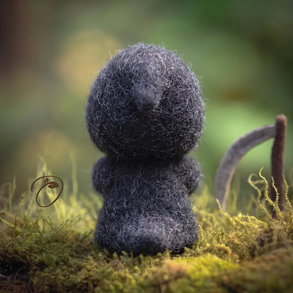 A small, fuzzy dark gray felt creature sits on mossy ground in a blurred outdoor setting with green and brown bokeh.