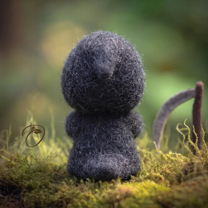 A small, fuzzy dark gray felt creature sits on mossy ground in a blurred outdoor setting with green and brown bokeh.