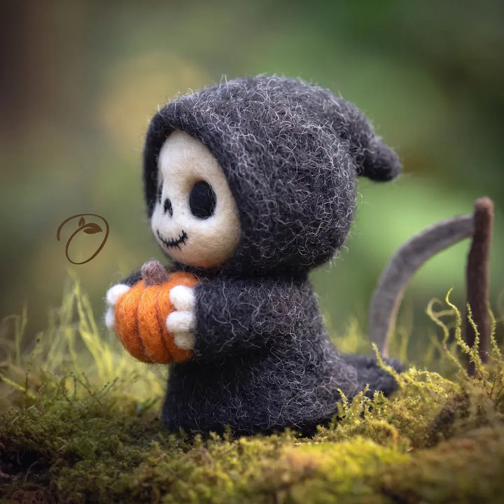 A small felted wool Halloween figurine of a skeleton in a dark gray hooded robe, holding an orange pumpkin and standing on mossy ground with a blurred forest background.
