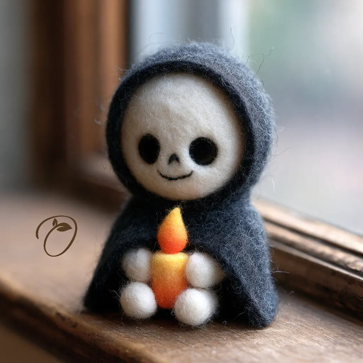 A small felt needle-art doll with a white skull face, black eyes, and a dark gray hooded body, sitting on a wooden windowsill and holding an orange candle.