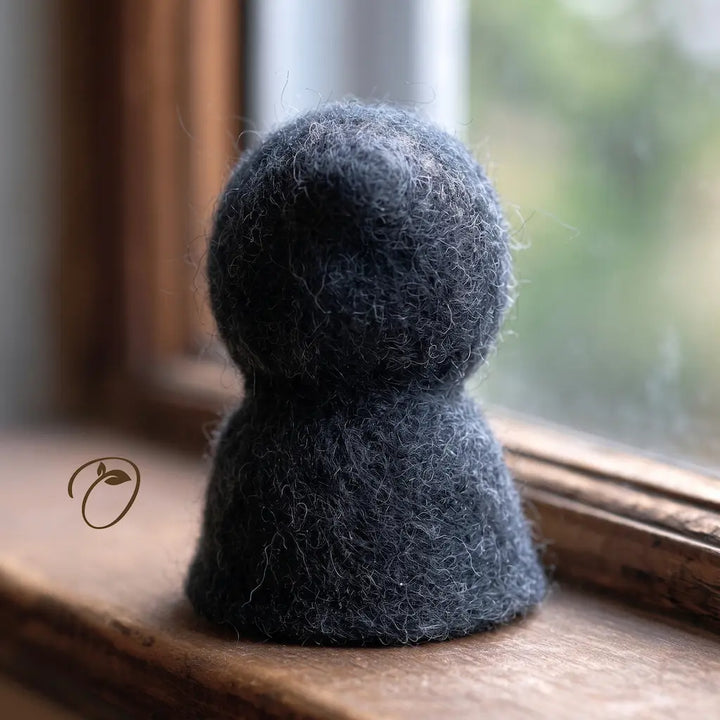 A dark charcoal gray felted wool decorative figurine shaped like a small gnome or character, resting on a wooden windowsill with a softly blurred window background.