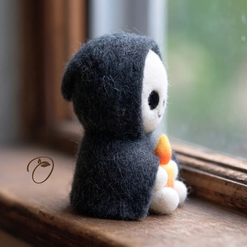A small felted plush toy with a dark charcoal gray hood and a white face sits on a wooden windowsill, holding a small orange and white flower. The background shows a softly blurred window view.