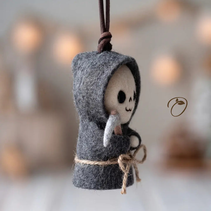 A small felted wool hanging ornament shaped like a hooded skeleton figure, with a dark gray hood, cream face, black eye details, a white hand, and a brown hanging cord. The background is softly blurred with warm bokeh lights.