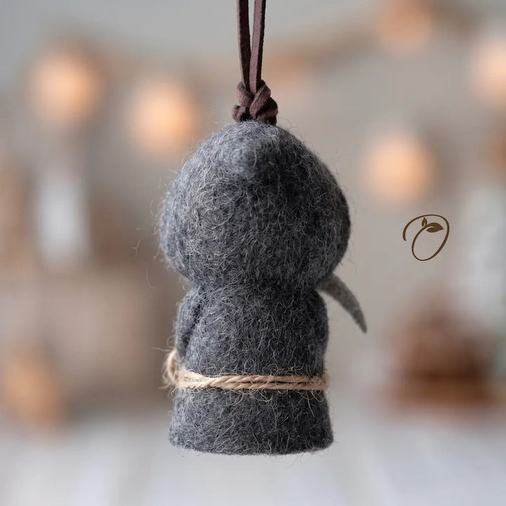 A small felted wool hanging ornament shaped like a seated figure, in dark charcoal gray with a natural twine wrap and a dark brown hanging loop, shown against a softly blurred warm neutral background.