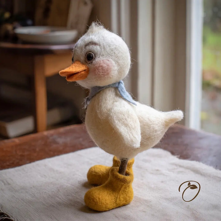 A needle-felted duckling figurine with a white wool body, orange beak, pink cheeks, a light blue scarf, and mustard yellow booties, standing on a textured fabric surface in a softly lit indoor setting.