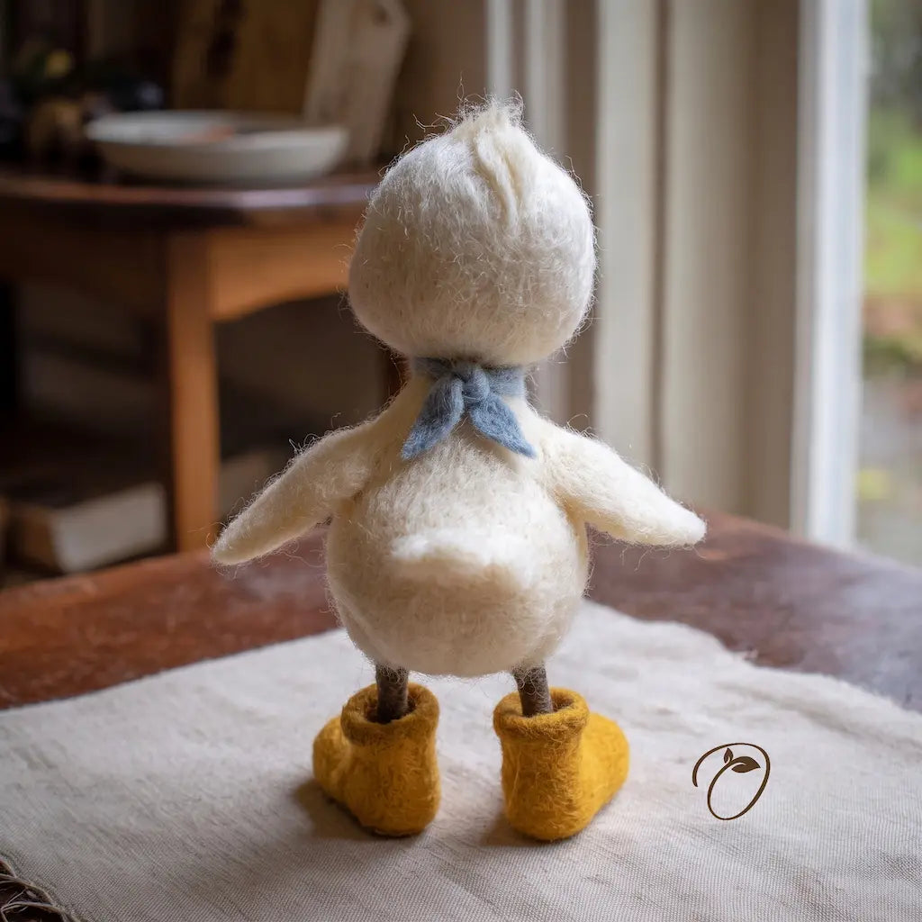 A small felted wool duck figurine shown from the back, standing on a light fabric surface indoors. The duck is cream-colored with a light blue bow and mustard yellow booties.