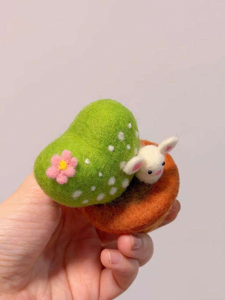 Hand holding a small felted toy with a green caterpillar, pink flower, and white rabbit on a plain background