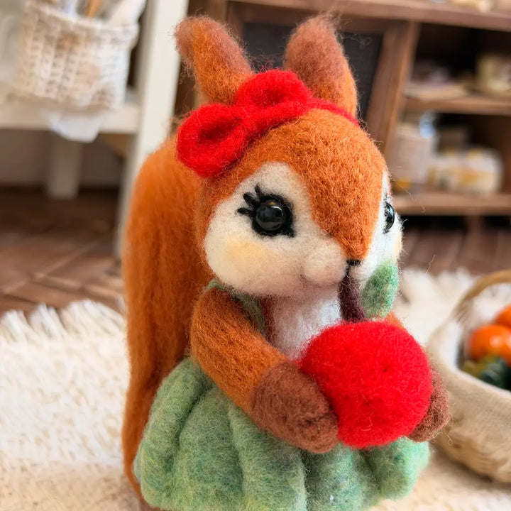 A needle-felted松鼠 toy holding a red apple, wearing a green dress and red bow, set in a cozy home environment.