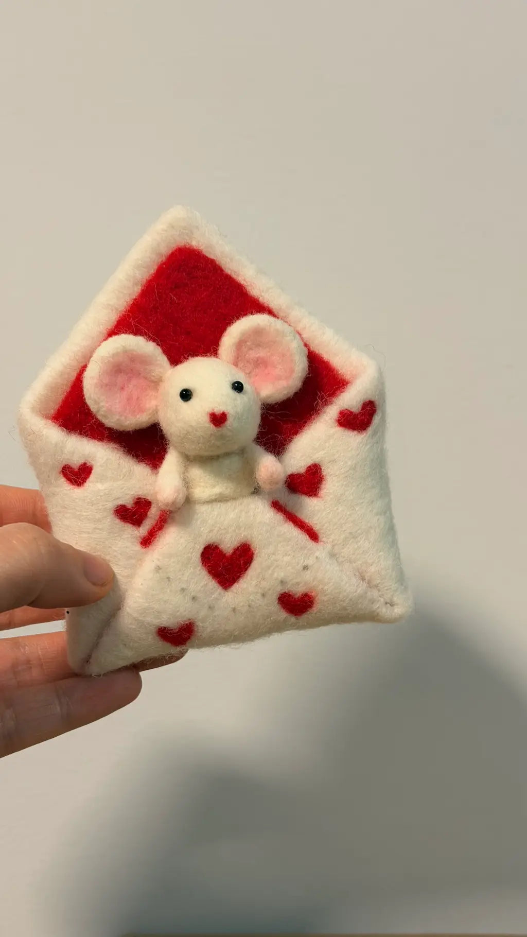 Small plush mouse toy inside a red envelope with heart patterns, held by a hand against a plain background.