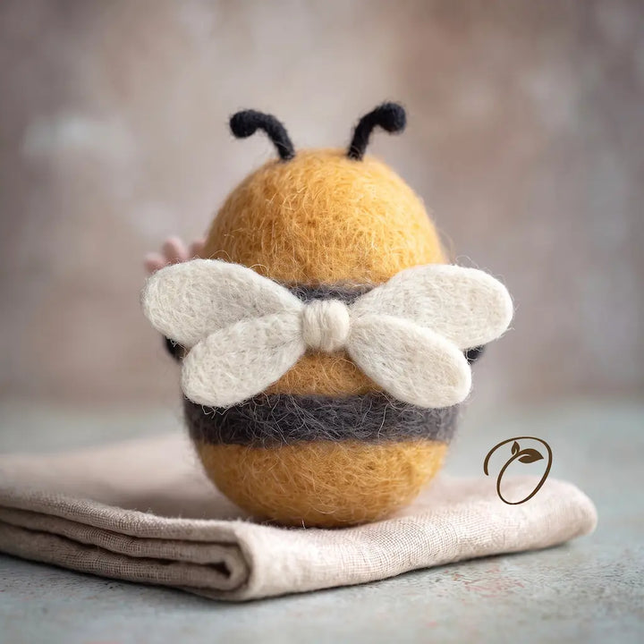 A small felt bee ornament or figurine with a yellow-orange body, black stripes, black antennae, and white wings, resting on folded beige fabric against a soft blurred neutral background.