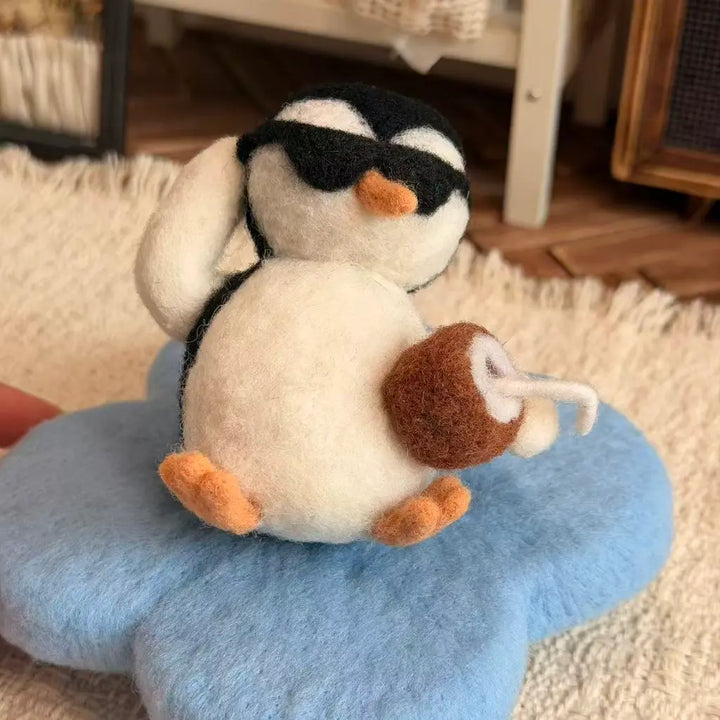 A small felt penguin toy wearing black sunglasses and holding a brown coconut drink, sitting on a blue flower-shaped cushion on a cream rug.