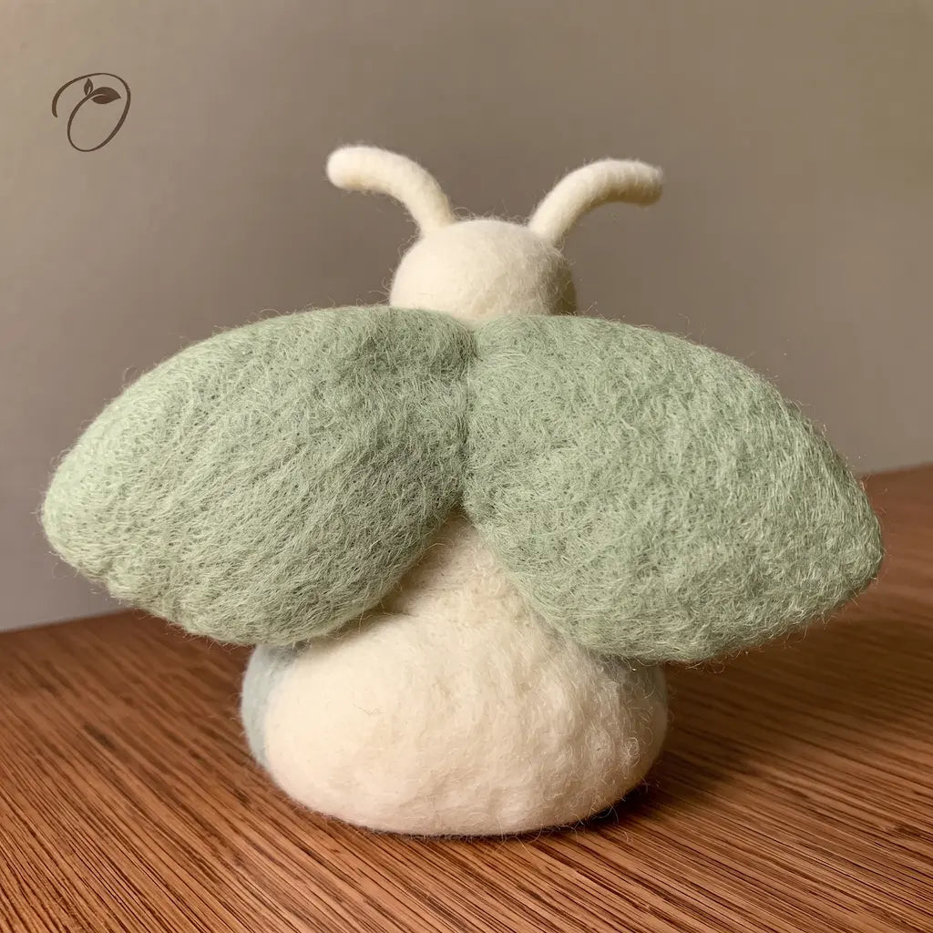 Back view of a wool felt moth with two broad sage-green wings, a rounded ivory body and two curved antennae.