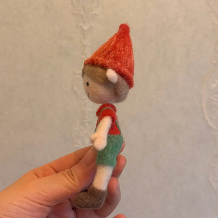 Hand holding a small elf figurine with a red hat and green pants against a plain background