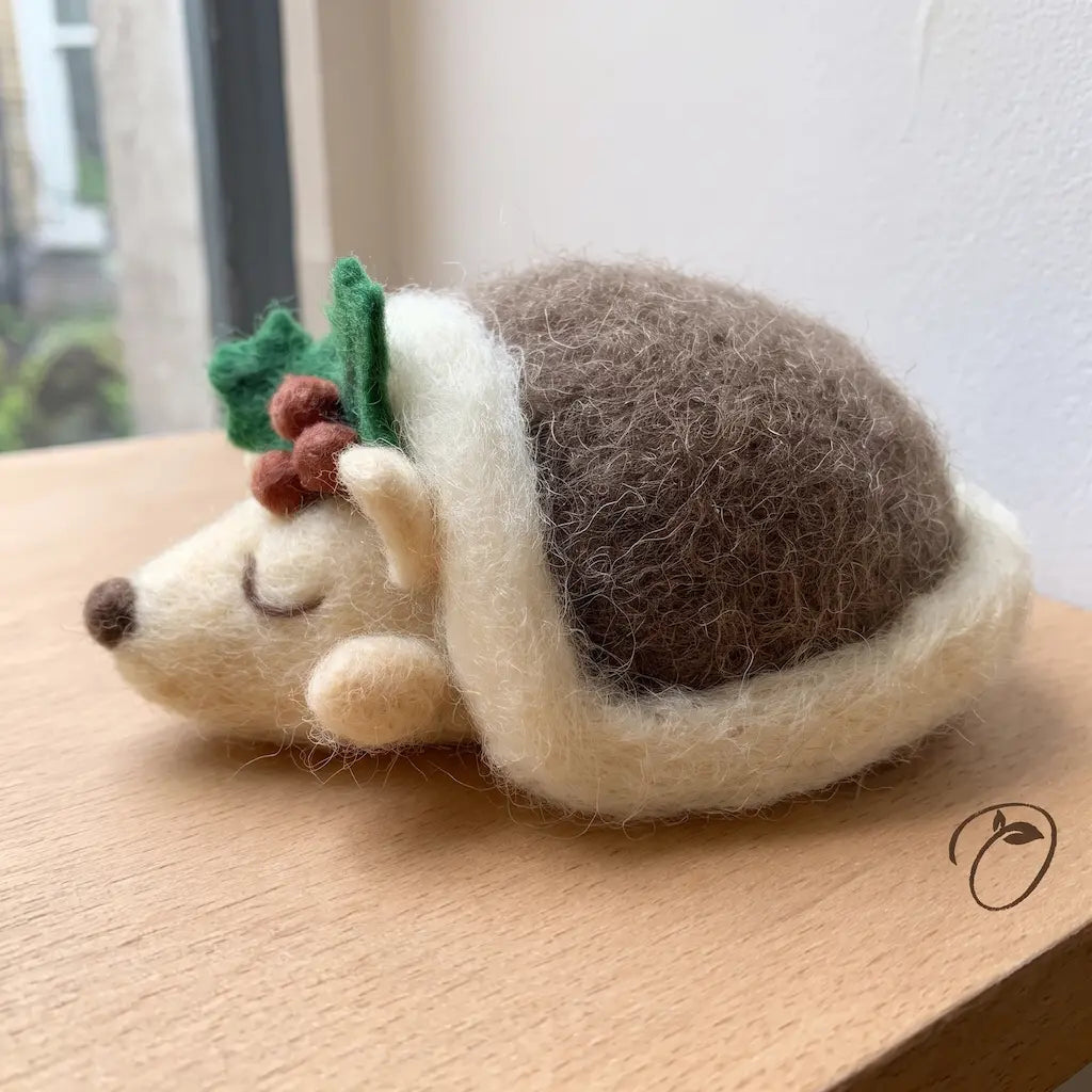Side view of a sleeping felt hedgehog with a domed brown back, cream face and trim, and holly leaves with red berries.