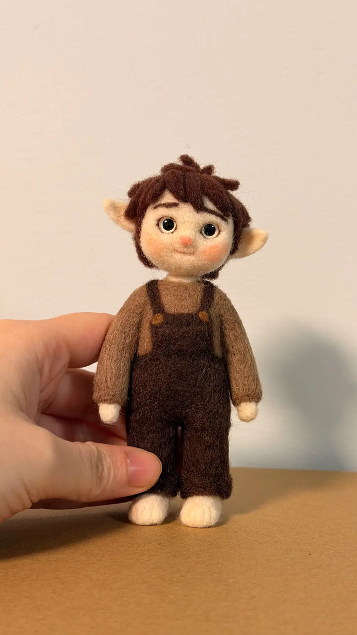 Small figurine of a character wearing brown overalls held by a hand on a beige surface.