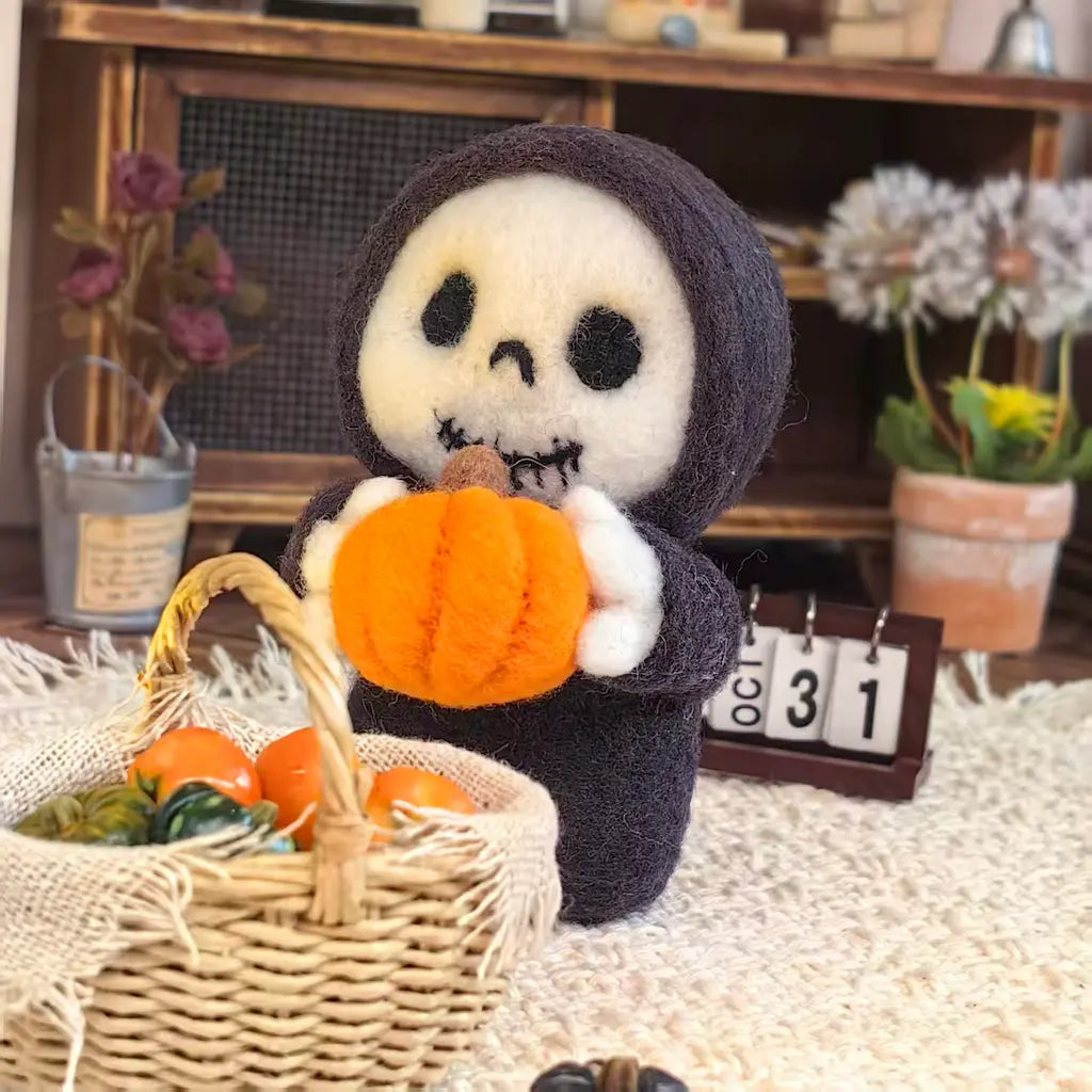 A felt needle art doll of a skeleton in a black hood holding an orange pumpkin, styled in a cozy autumn scene with a wicker basket, pumpkins, and a wooden calendar.