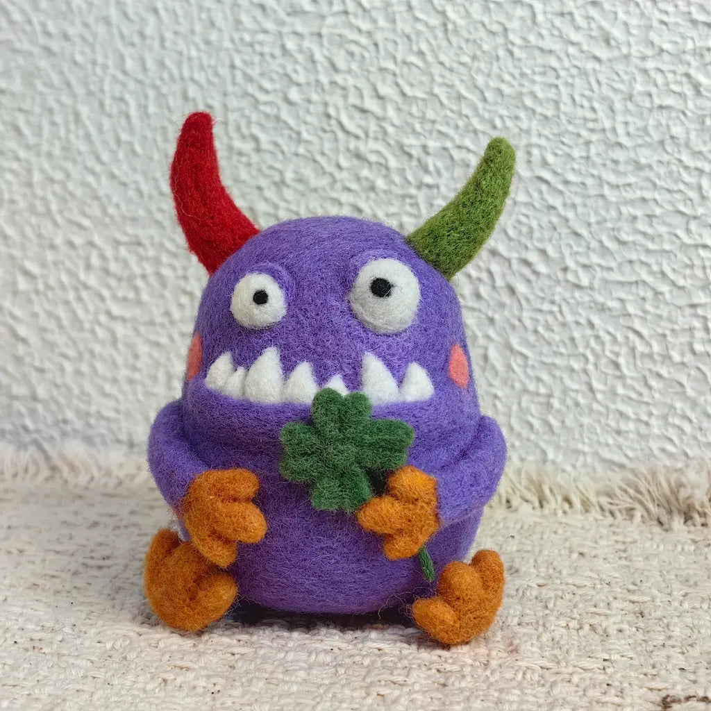 Front view of the Clover Monster needle-felted wool figurine with wide eyes, white pointed teeth, rosy cheeks and a green four-leaf clover held in its orange hands.