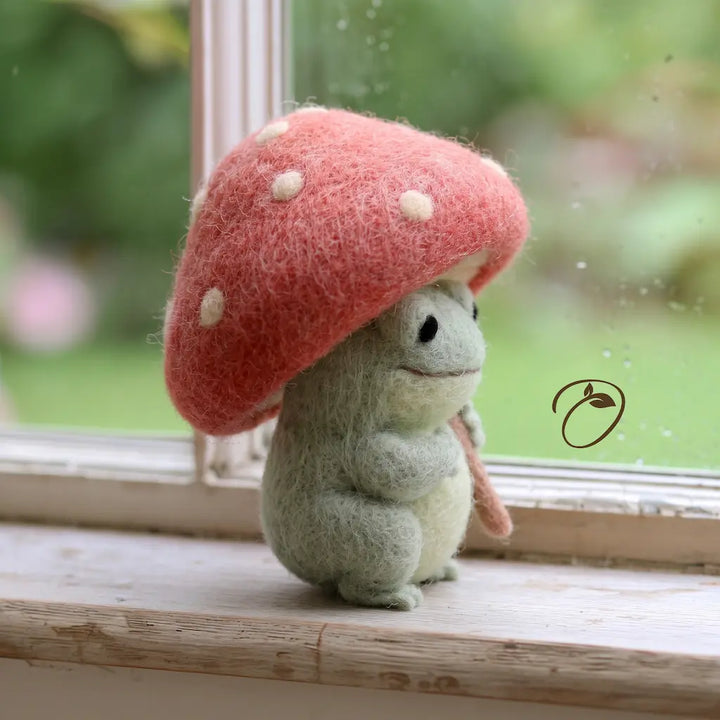 Side view of a smiling felt frog beneath a pink mushroom umbrella, with a pale belly and rounded paws