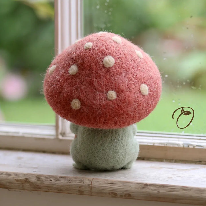 Back view of a green felt frog beneath a broad pink mushroom cap dotted with cream spots