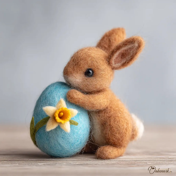 Felted bunny holding a blue egg with a yellow flower on a wooden surface.