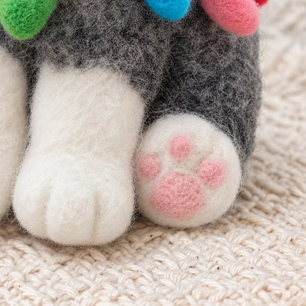 Close-up of a handmade Old English Sheepdog felt figurine showing its fluffy white paws, pink wool paw pads, textured gray coat, and colorful felt Christmas lights.