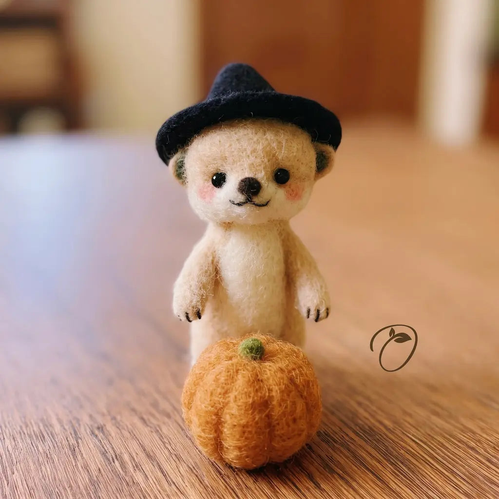 A small needle-felted teddy bear wearing a black witch hat, standing on a wooden surface next to a small orange pumpkin. The background is a softly blurred indoor setting.