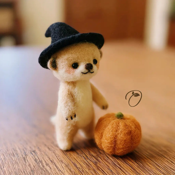A small felt teddy bear figurine wearing a black witch hat, standing on a wooden surface next to a small orange pumpkin. The background is a softly blurred indoor setting.