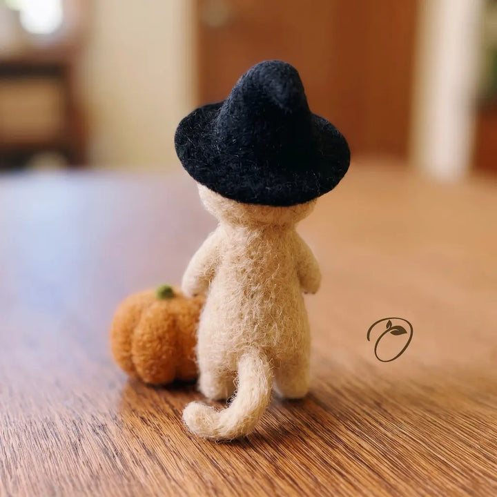A small beige felt animal figure wearing a black hat, shown from the back on a wooden surface with a small orange pumpkin beside it. The background is softly blurred indoor decor.